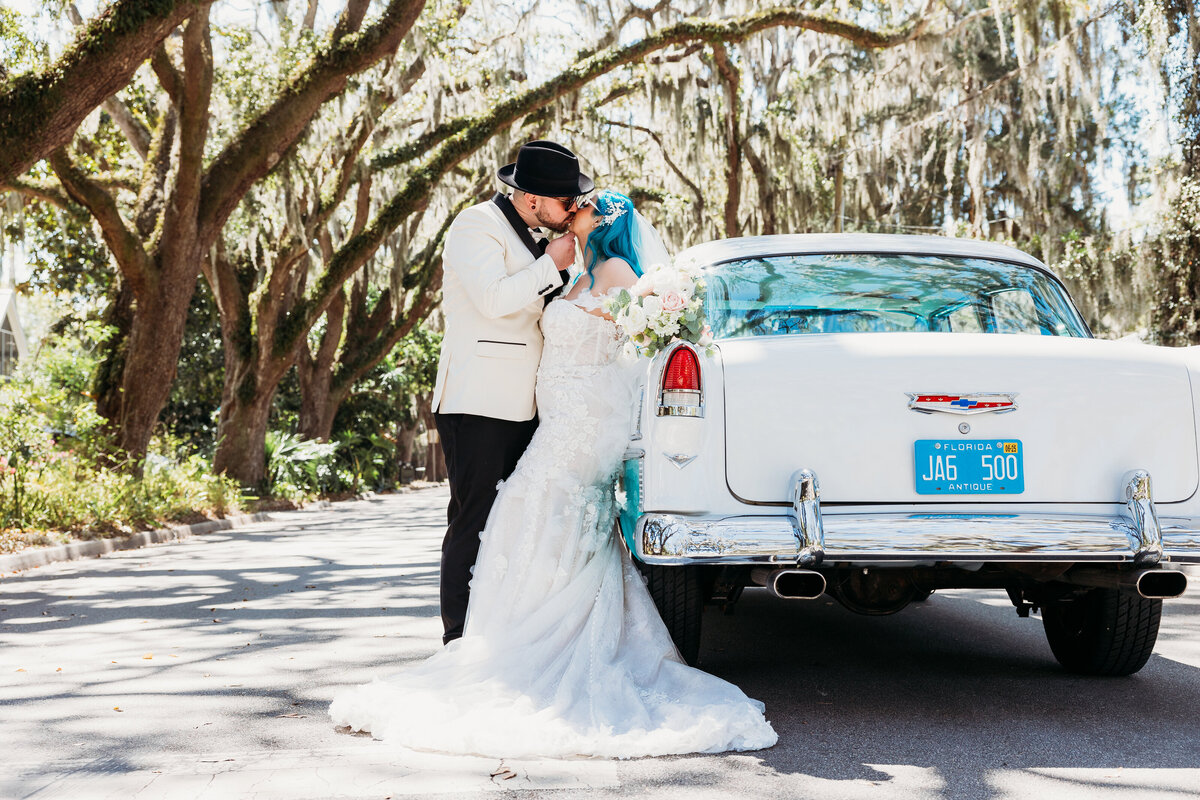 Groom in fedora and bride with blue hair share a kiss by a vintage white car under moss-draped oaks in downtown St. Augustine.