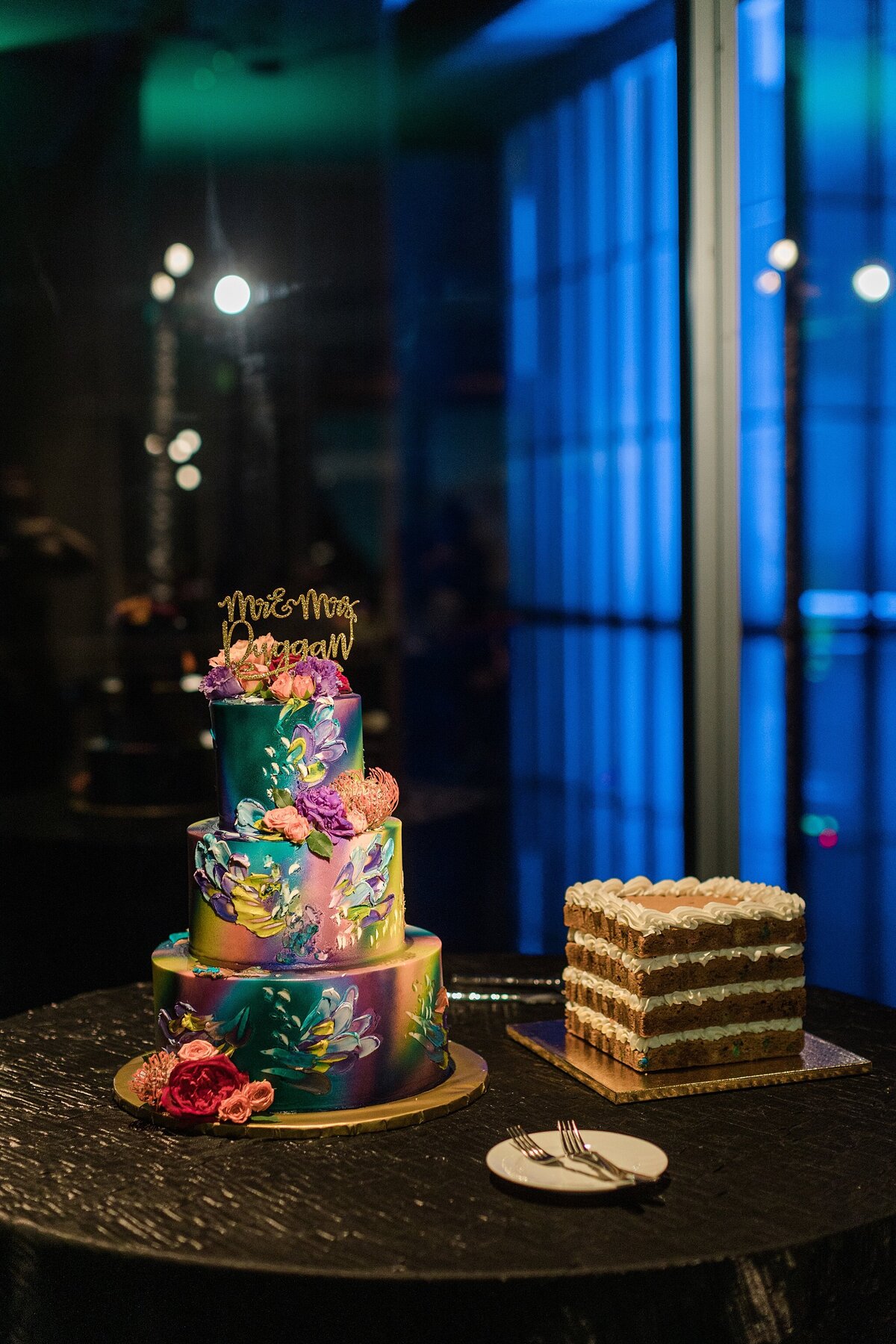 A detail shot of the cake table at a wedding reception at the Modern Art Museum of Fort Worth in Fort Worth, Texas. The cake on the left is a wildly colorful, three tiered cake with colorful flowers and floral decoration with a gold topper show the bride and groom's new last name. The cake on the right is a four tiered cookie cake with white icing trimming the edges.