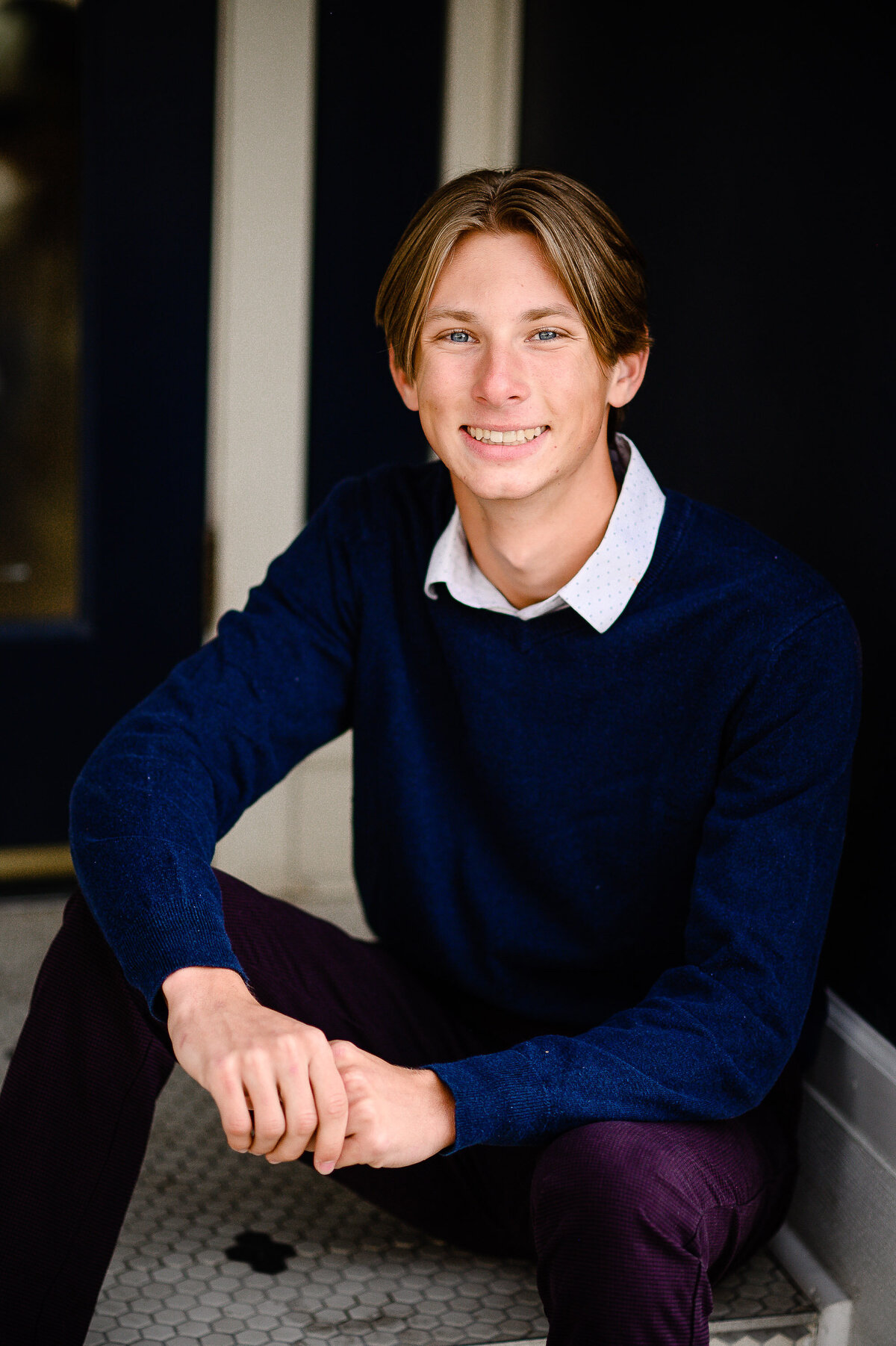 senior picture outfits for guys with young man in purple pants and a blue sweater sitting on the floor and leaning against a wooden wall  photographed by denver senior photographer