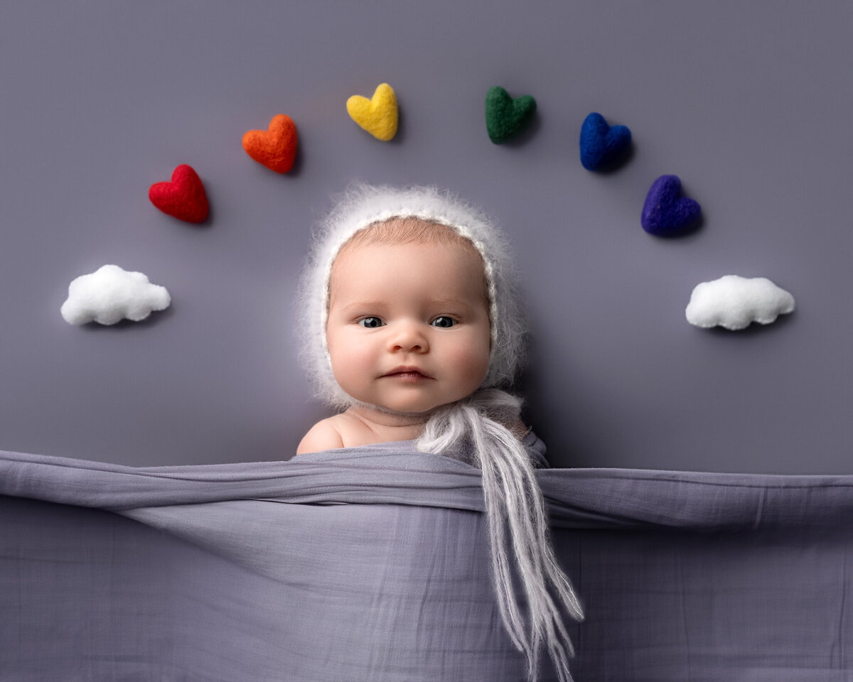 Rainbow baby with white knit bonnet surrounded by colorful felt hearts and clouds on a purple background in Austin and surrounding cities