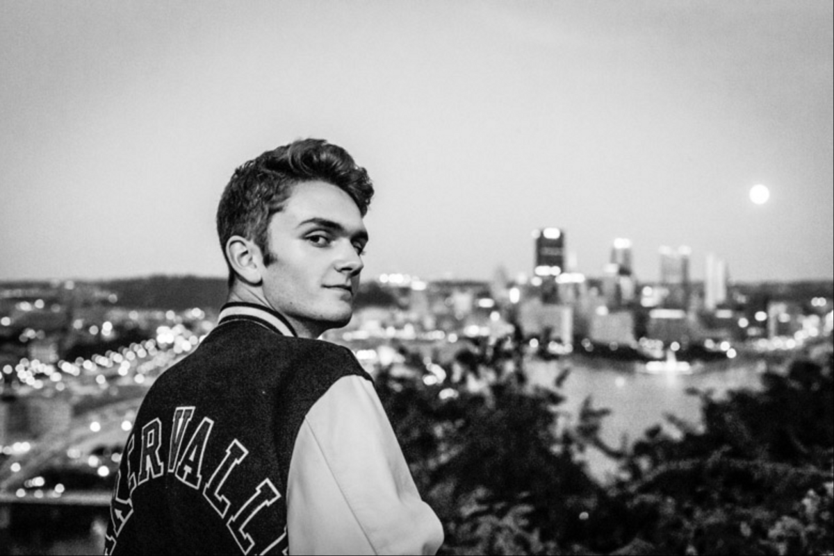A contrast the black-and-white image of a boy wearing A letterman's jacket looking back at the camera with the city of Pittsburgh skyline in the moon in the backdrop.