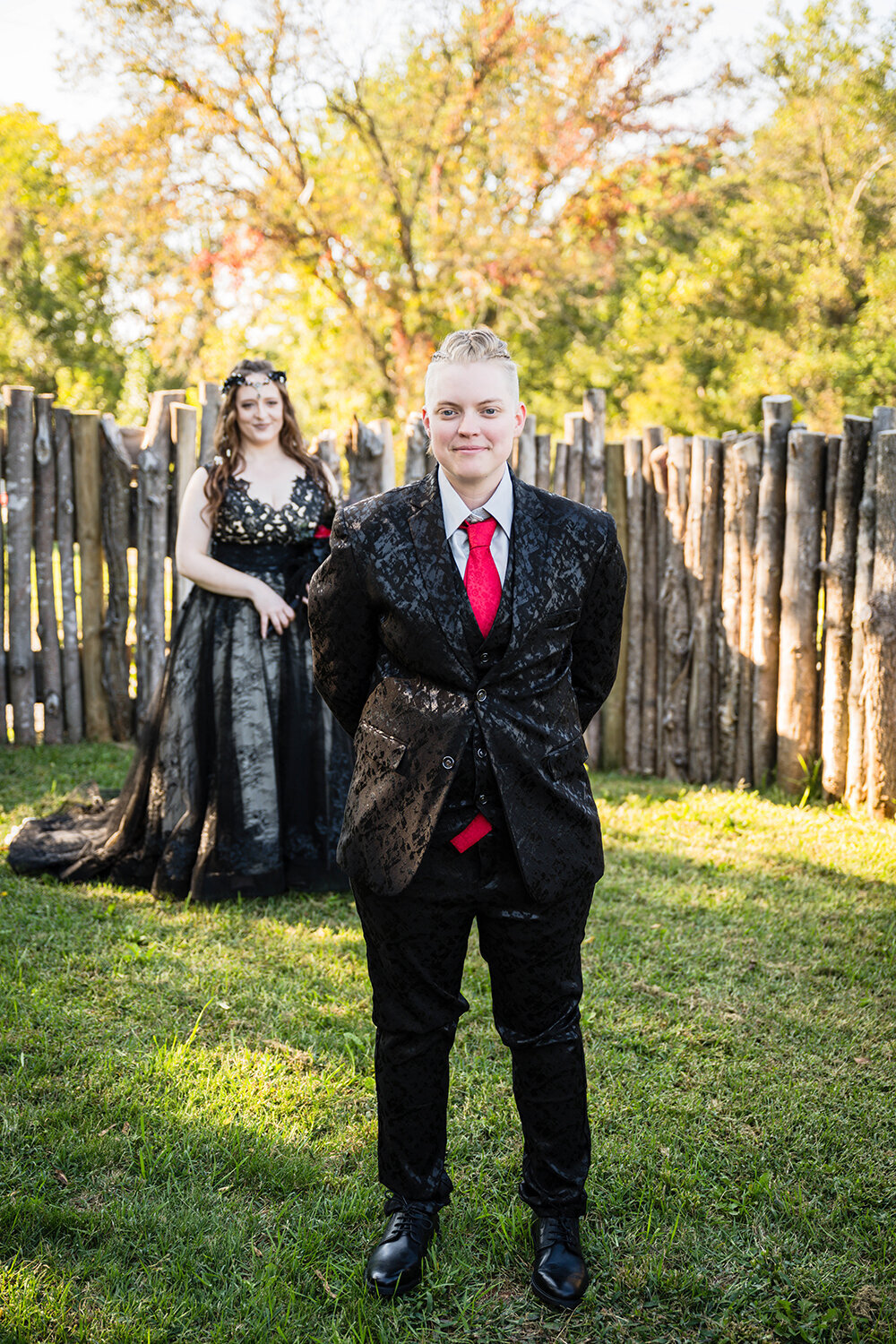 Two LGBTQ+ marriers stand in the backyard of an Airbnb they rented in Roanoke, Virginia on their elopement. One marrier, dressed in a black floral suit, stands in front of another marrier, who is wearing a black dress. The marrier in the background looks at the back of the other’s head while the marrier in front grins waiting.
