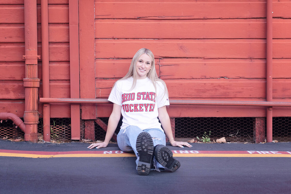 A person with long blonde hair sits on the ground, leaning against a red wooden wall. They are wearing a white Ohio State Buckeyes T-shirt and jeans, with legs extended and crossed at the ankles.