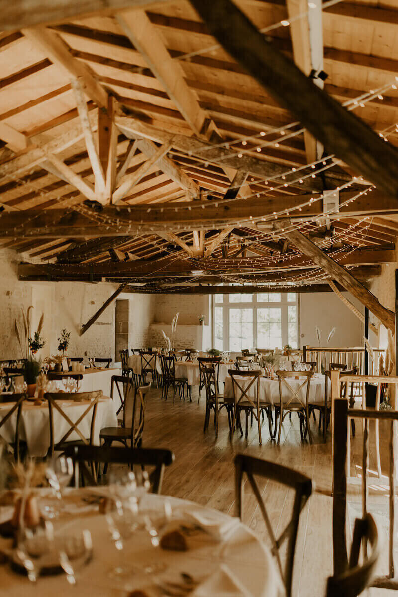 Salle de cérémonie de mariage avec chaises et poutres en bois et guirlandes de lumières au plafond. Photo prise par Laura Termeau photographie.