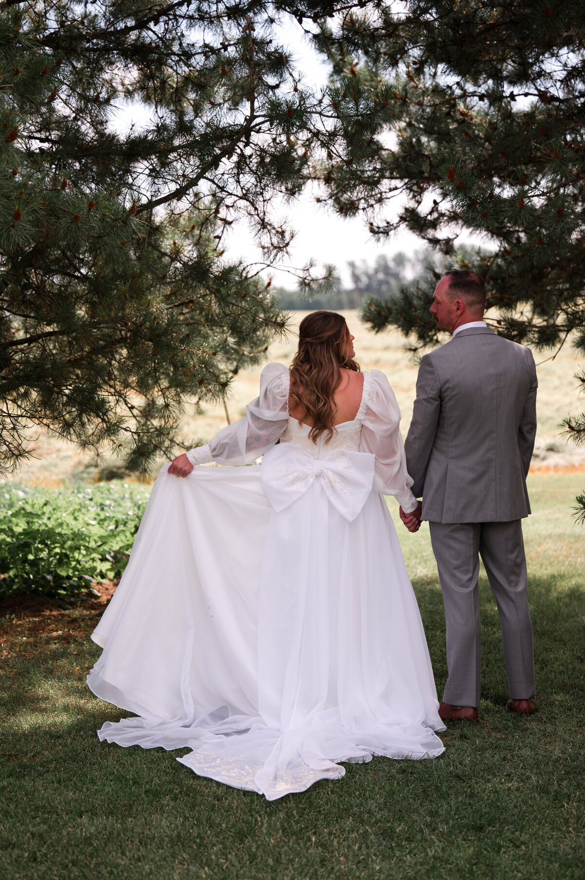 bride and groom pose for wedding photos surrounded by lush greenery, bride is holding her dress out so that it flows in the wind