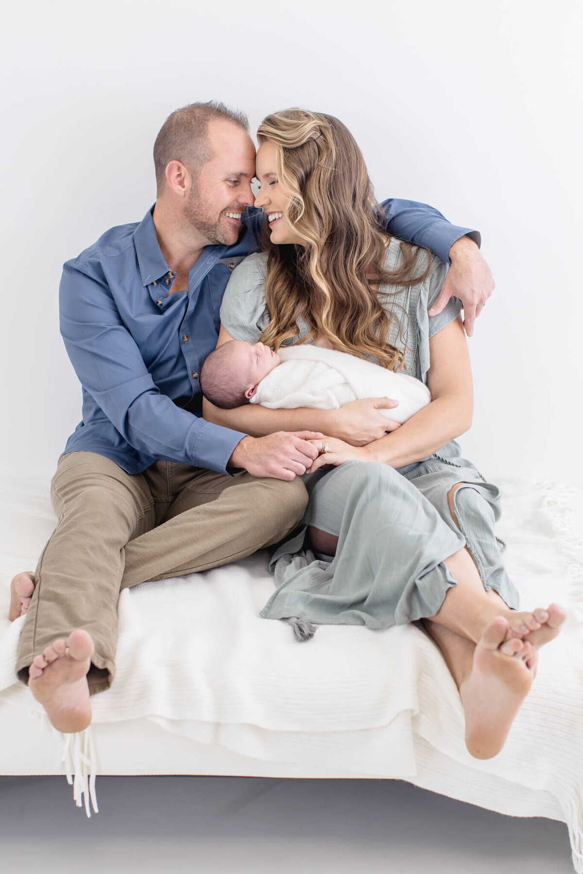 A couple sits closely on a bed, gazing at each other affectionately. They hold a baby wrapped in a white blanket. The man wears a blue shirt and khaki pants, while the woman is dressed in a light blue dress. The setting is bright and serene.
