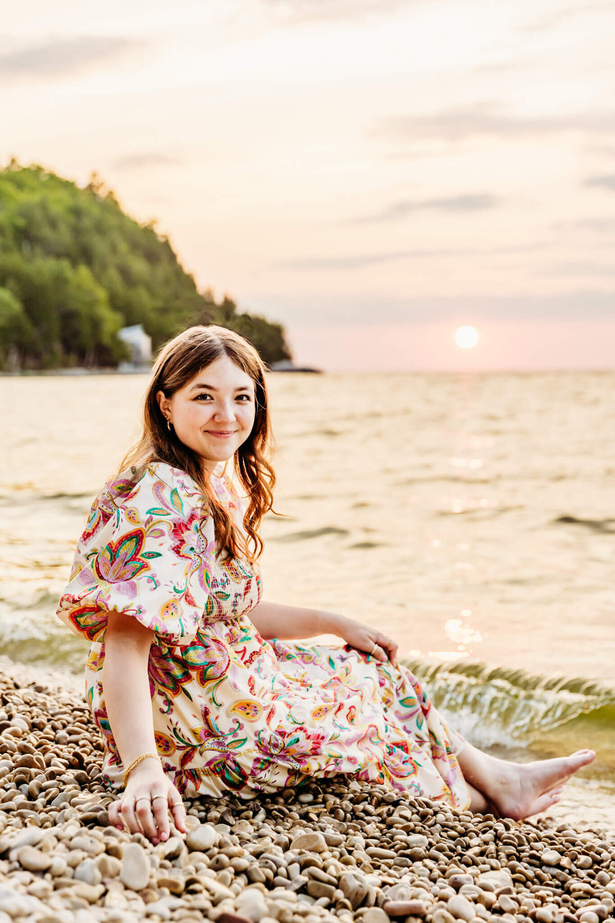 gorgeous teen girl in a colorful patterned dress sitting on a white stone beach and reaching back and smiling captured by senior photographer Ashley Kalbus