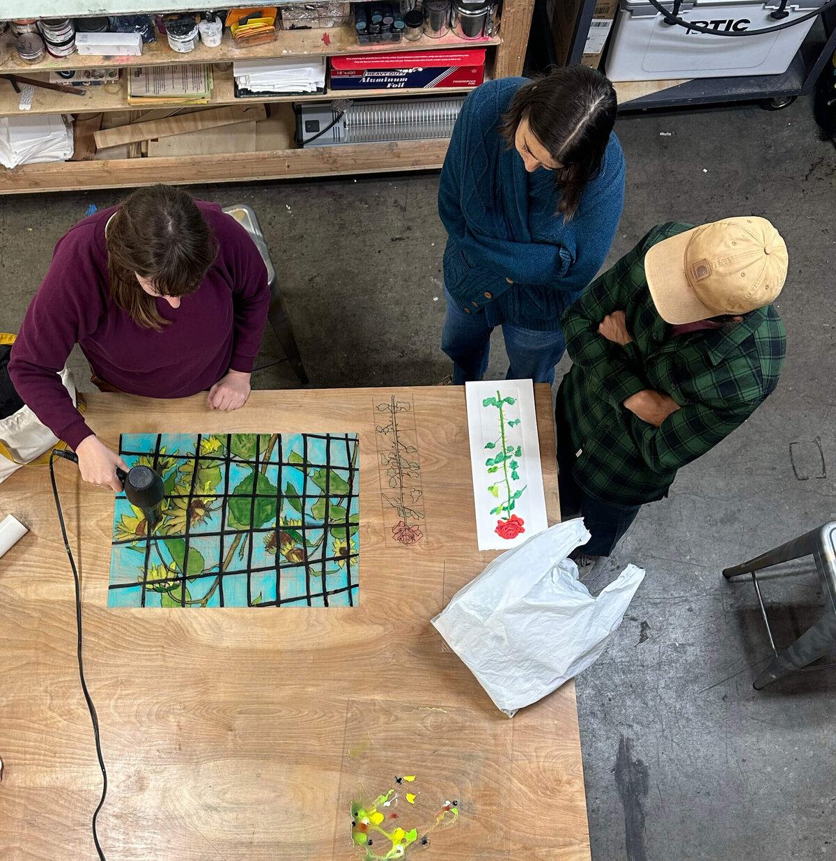 Overhead view of a monoprint demo in progress, with a sunflower design on plexiglass and participants watching the technique unfold.