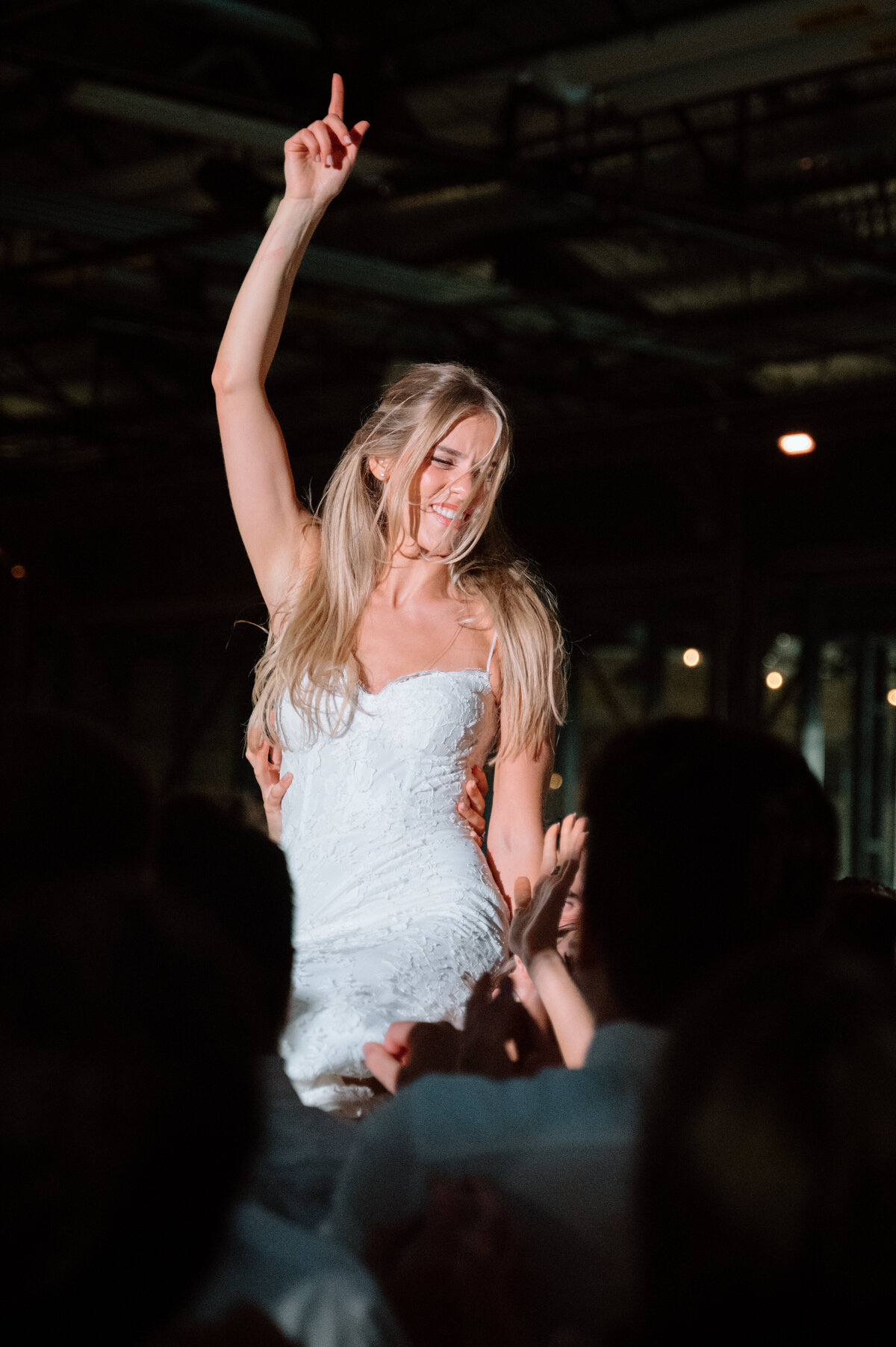 Bride smiling with arm raised while lifted in a chair during celebration.