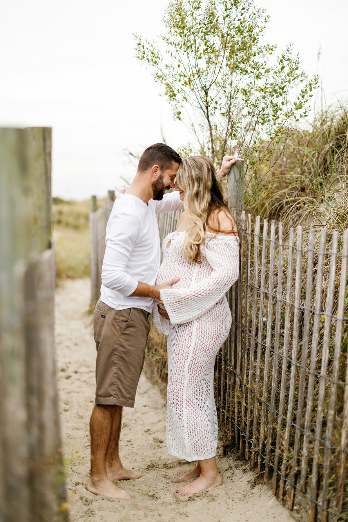 Expectant couple facing each other closely by beach fence in Wilmington, jointly cradling baby bump