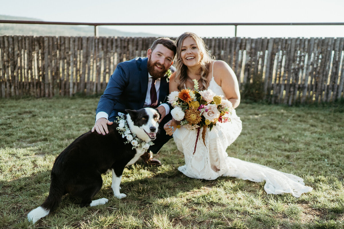 Bride and groom pose with their dog and a fall colored bouquet on their wedding day.