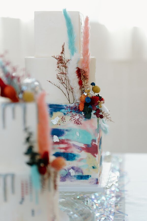 Photo focuses past part of one cake onto part of the middle cake. Middle cake is three tiers with multicolored watercolor swatches on the bottom square tier. There's accents of silver leaf, bright dried florals, and mini disco balls.