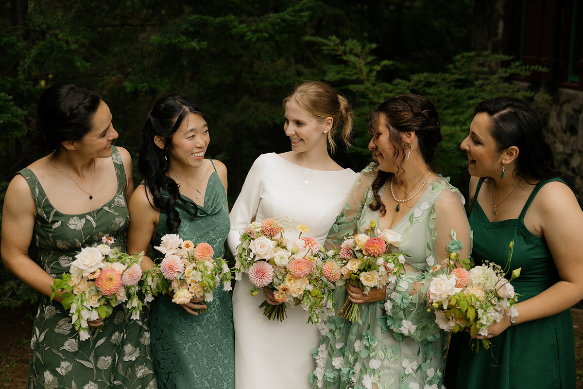 Bridesmaids in green dresses surround a bride in, holding bouquets of peach flowers at a Montreal wedding.