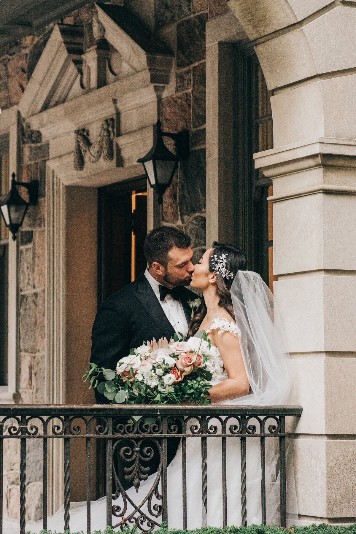Bride and groom kiss on balcony holding gorgeous bouquet of white and pink florals