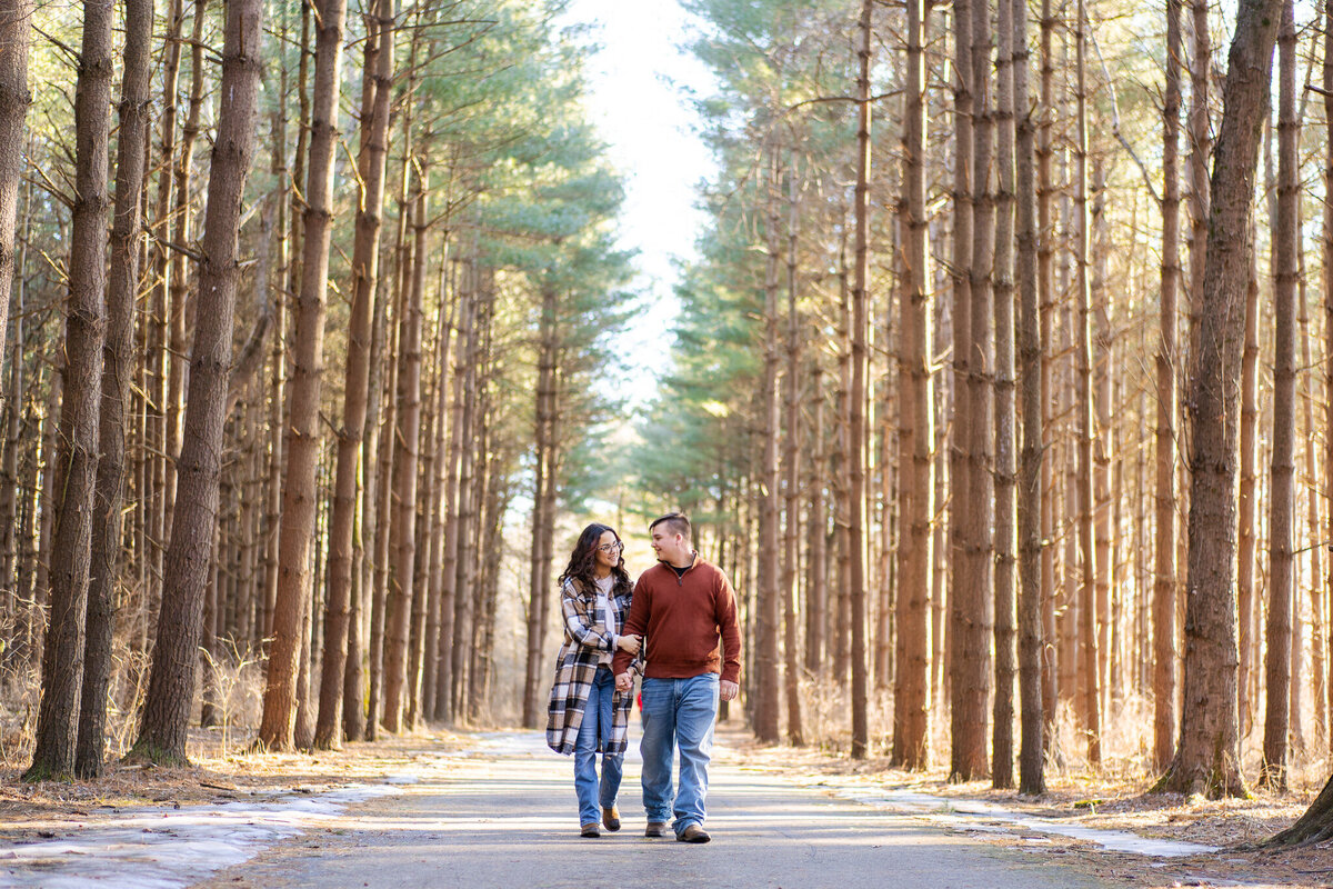 An engaged couple looking at each other and holding hands as they walk through a wintery path of tall pine trees - Walnut Woods Tall Pines in Groveport, Ohio.