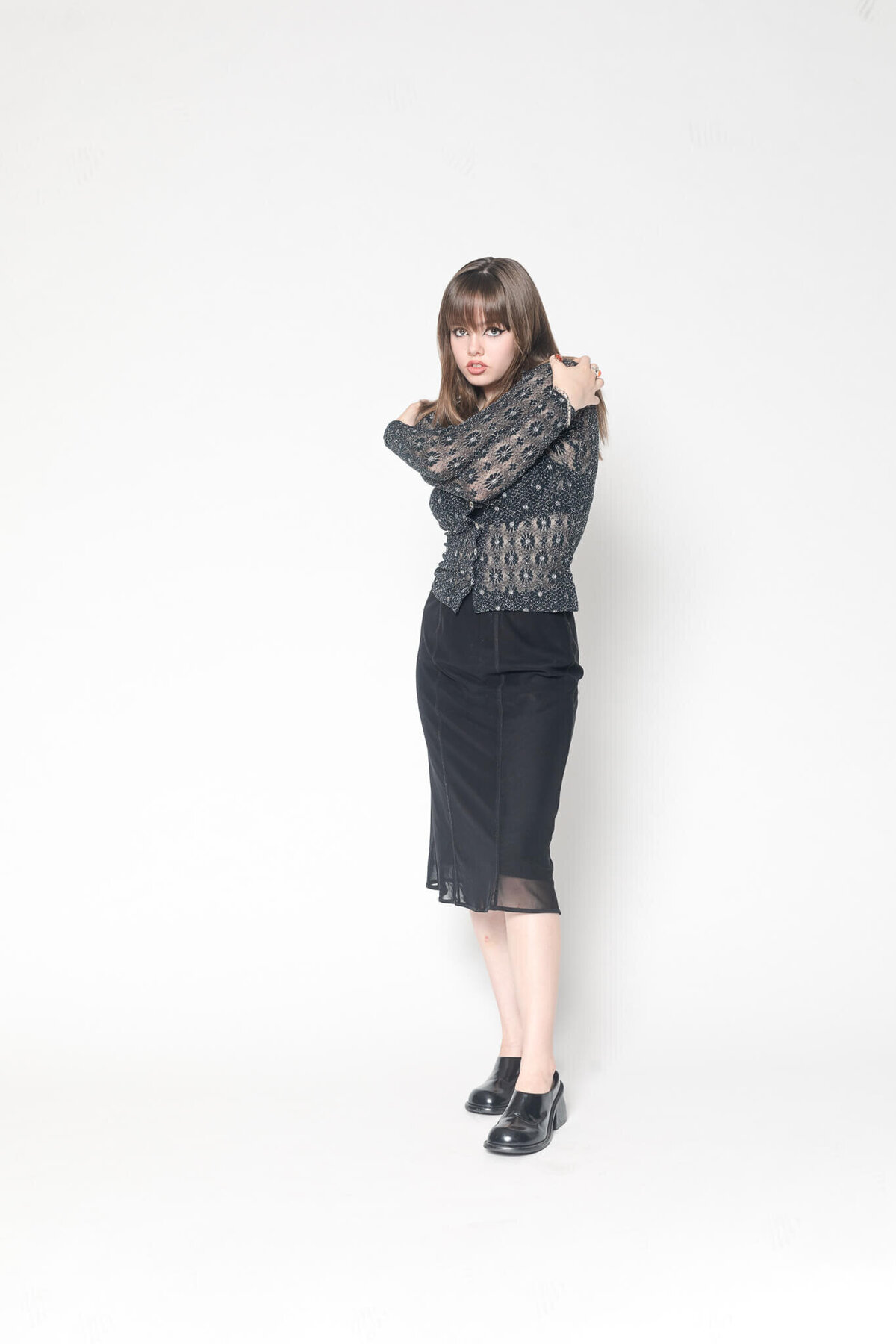 A person with long hair stands against a plain background, wearing a patterned, long-sleeved top and a black skirt. They have their arms crossed over their chest and are wearing black shoes.