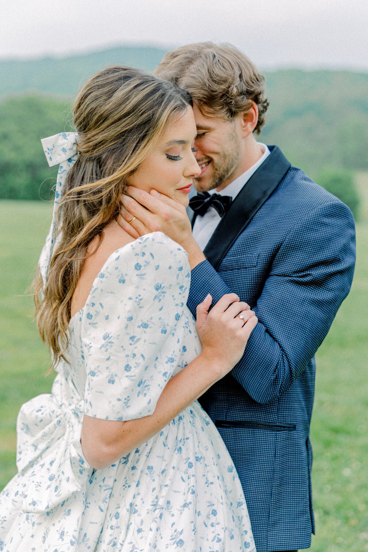 Ivy Rose Barn | A romantic bride and groom embrace in a sunlit field, surrounded by rolling green hills. The bride wears a vintage-inspired floral gown with puff sleeves and a bow, while the groom, in a navy tuxedo, holds her close. Captured by a Richmond wedding film photographer.