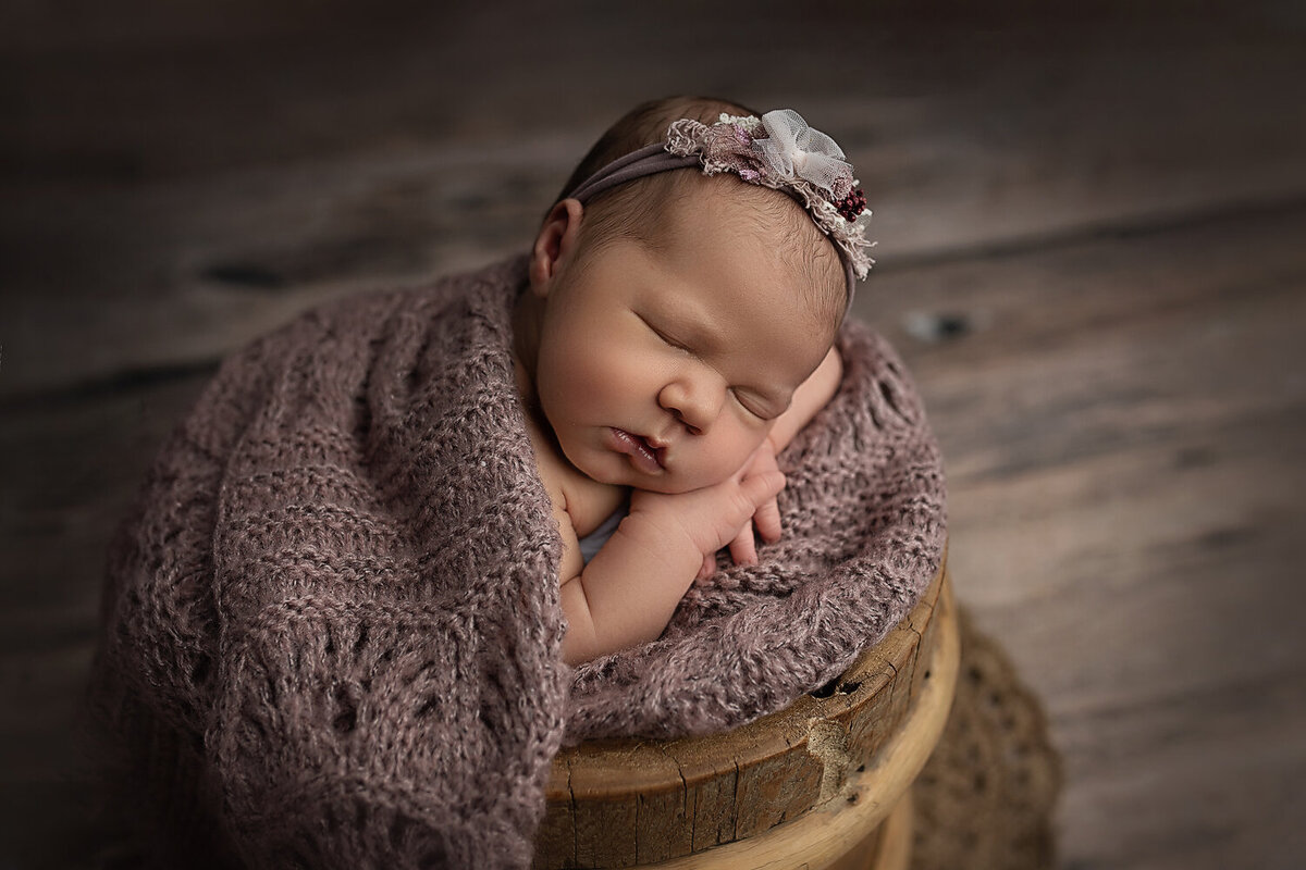 A New Orleans Newborn Photographer posed a sleeping newborn girl in a bucket wrapped in a purple knit blanket
