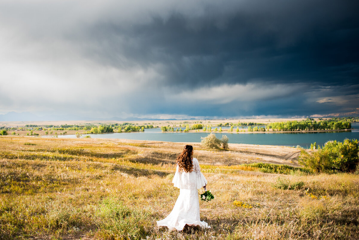 A wide angle shot of a bride in a field with a lake in the background and stormy sky. The bride walks away from the camera.