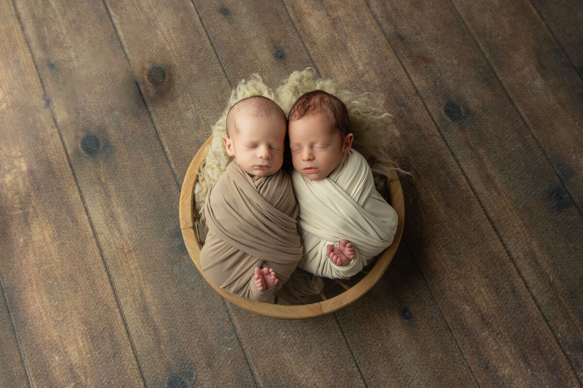 Two newborn twins lie side by side in a circular basket lined with soft materials. One twin is wrapped in a light brown swaddle, while the other is in a cream swaddle. They have calm expressions, and their tiny hands are partially visible. The background features a textured wooden floor that adds warmth to the overall scene, emphasizing the gentle and serene atmosphere of the moment.