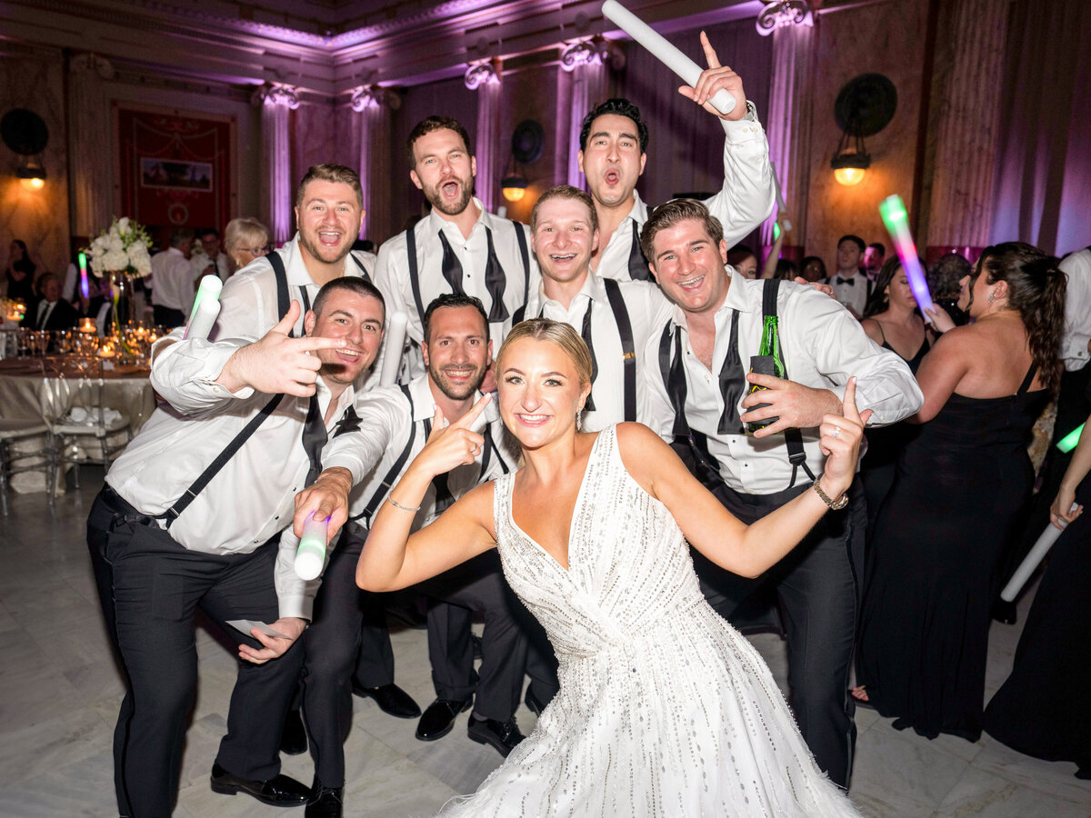 A bride in a white gown poses energetically with a group of eight men at an indoor wedding reception. The men are dressed in formal attire and hold glow sticks, with purple lighting in the background creating a festive atmosphere.