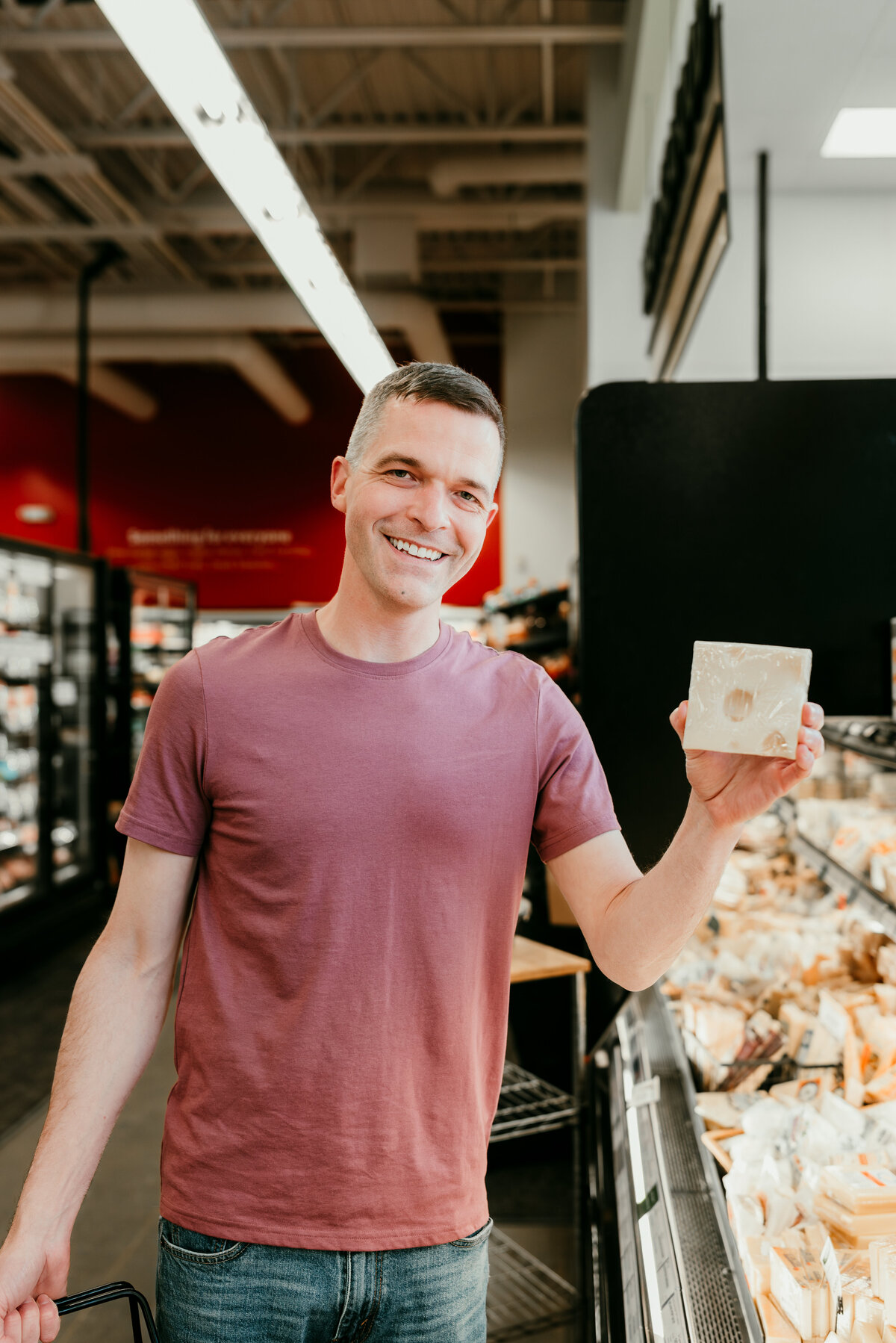 Man in a casual pink t-shirt holding a drink in a grocery store aisle, showcasing a fun and approachable personality.