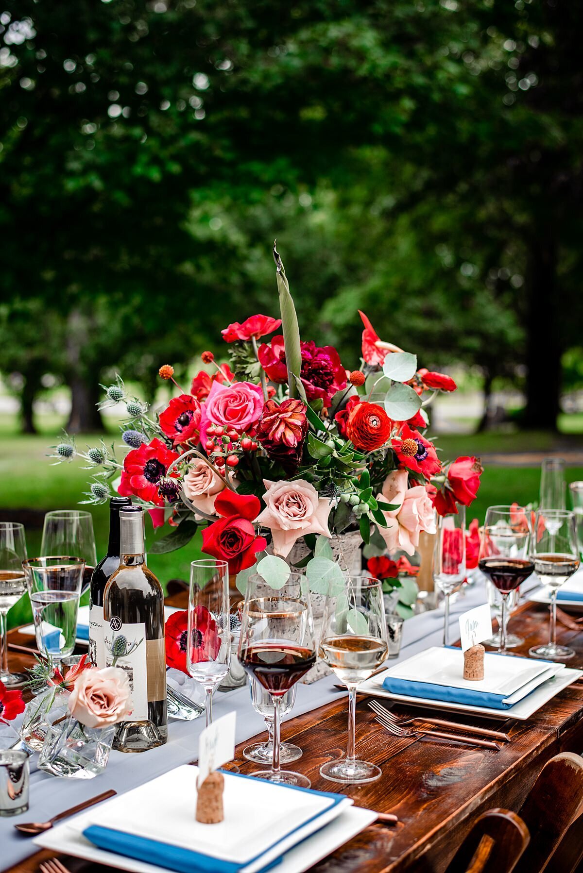 A farm table set with a large centerpiece of red roses, blush roses, red anthurium, red ranunculus, red anemones, red and pink protea, blue thistle, hypericum berries, red cascading dianthus and assorted greenery. Square glass bud vases of blush and red roses, silver and gold votive candles and wine cork seating card stands decorate the table along side bottles of Arrington Vineyard wines. Square white dinner plates are layered with a teal linen napkin wrapped salad plate and a square white dessert plate with matte copper flatware and delicate stemmed red and white wine glasses and footed cut crystal water glasses with a gold rim at a southern summer wedding at Arrington Vineyards.