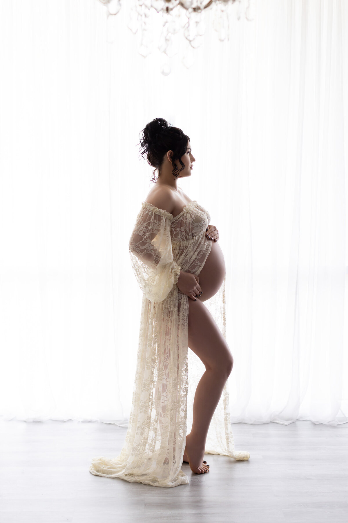 A stunning side-profile maternity portrait showcasing a woman standing elegantly in a sheer, lace gown with flowing sleeves. Soft natural light filters through white curtains, creating a bright, ethereal backdrop. This luminous portrait captures a moment of peaceful anticipation, highlighting the graceful curve of her silhouette. Captured by Lindsay Walden Photography, Dallas maternity photographer.