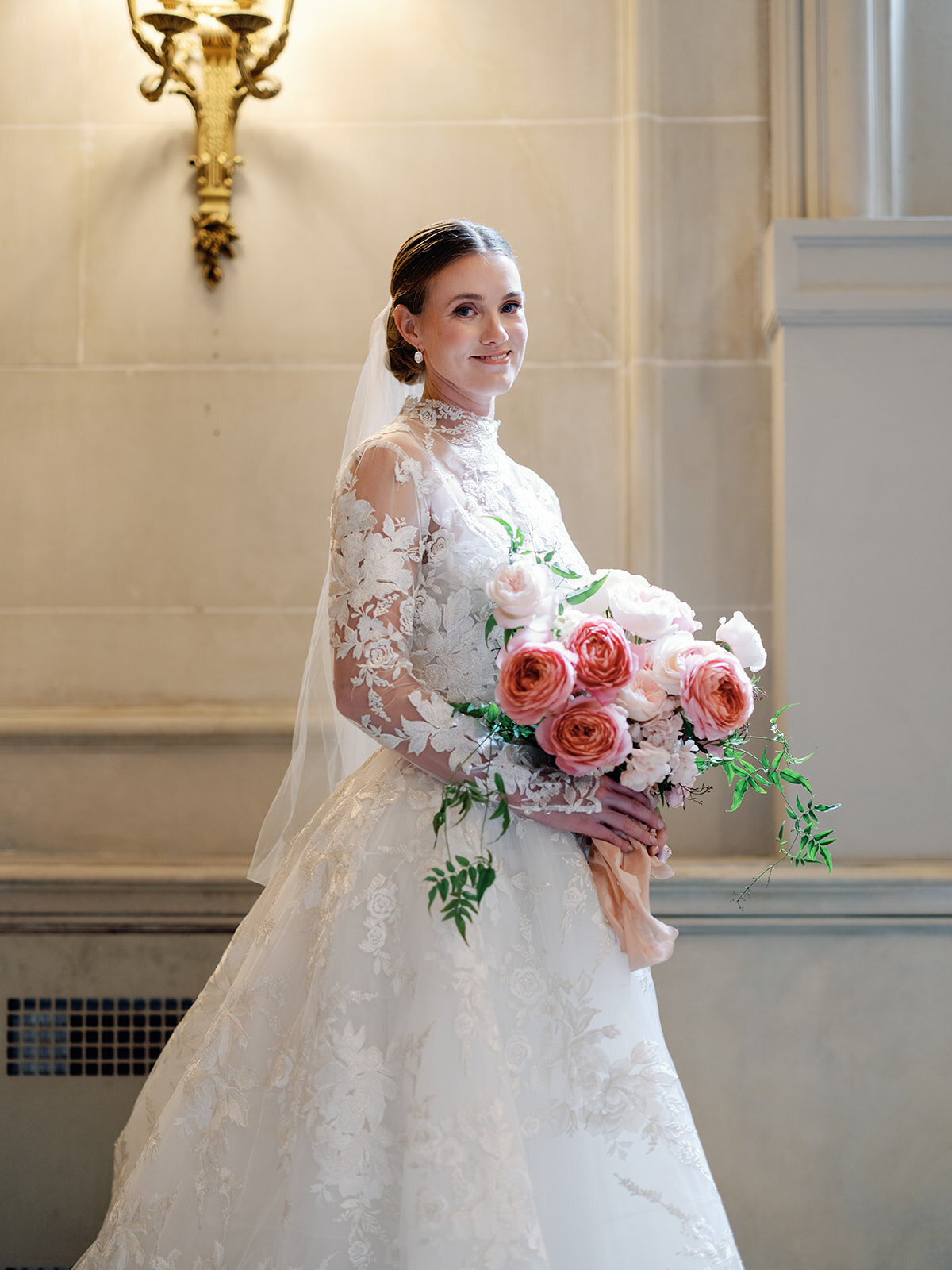 A bride stands by window light holding her  blush pink bouquet