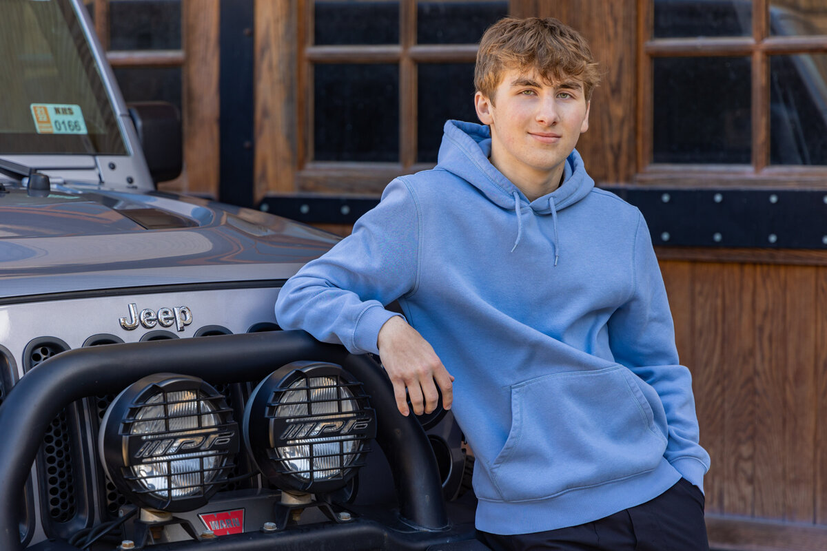 A male high school senior is posing with is right arm resting on the hood of his Jeep.  There are large wooden doors behind the truck.  The young man is wearing a blue hoodie sweatshirt