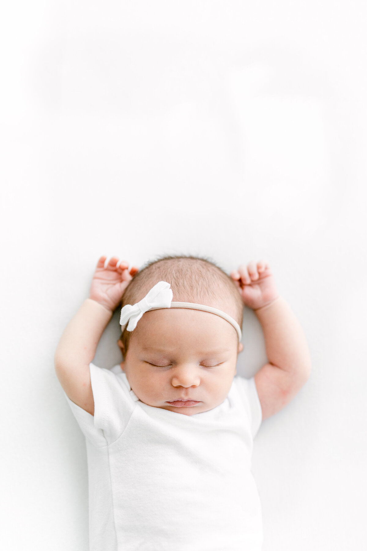 Headshot of a newborn baby against an all-white blanket with their arms up above their head and eyes closed