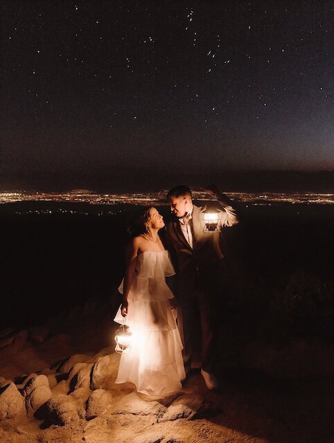A couple holding lanterns and standing under a starry sky at night, with city lights visible in the distance at Joshua Tree National Park.