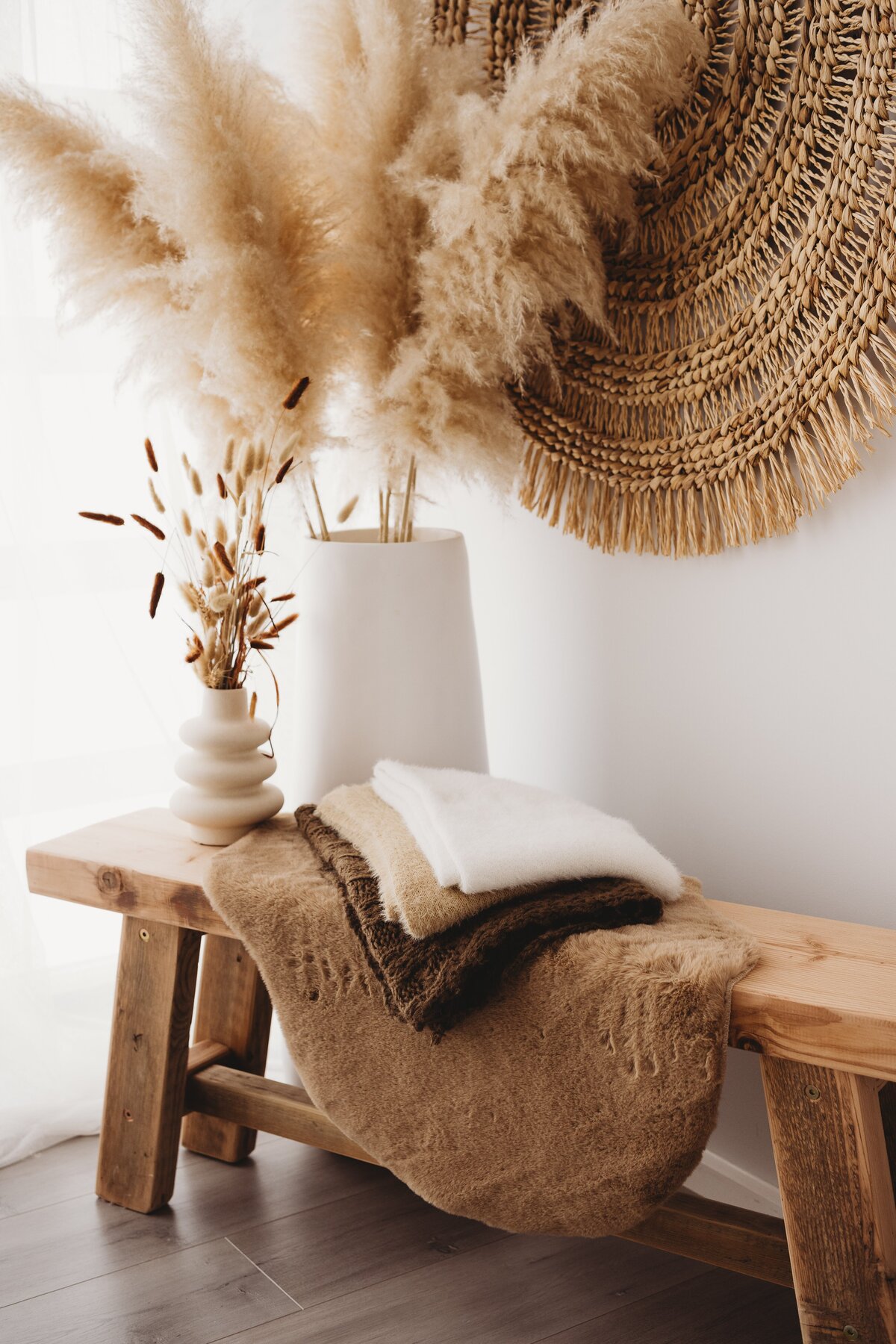 A wooden bench with a woven mat, stacked folded blankets, and decorative vases holding pampas grass and dried plants is situated in a minimalist room—a perfect setting for NSW family photographer Wollongong motherhood sessions. A woven wall decor hangs above.