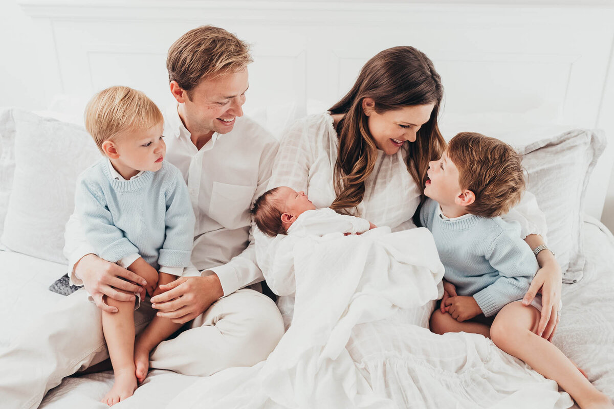 a family of five sit on a bed together cuddling and smiling at each other