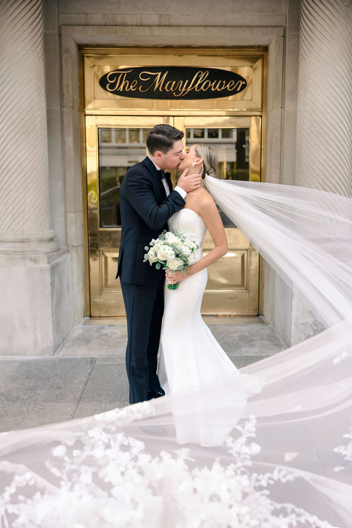 A bride and groom share a kiss in front of a building with The Mayflower sign. The bride wears a white gown and veil, holding a bouquet, while the groom wears a black suit. The long veil flows elegantly in the foreground.