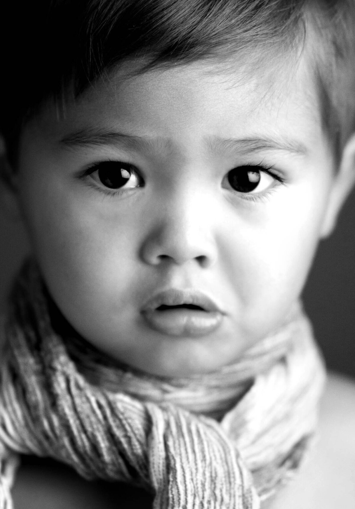 A black and white close-up of a young child with wide eyes and slightly open lips, wearing a textured scarf. The expression appears curious and thoughtful.