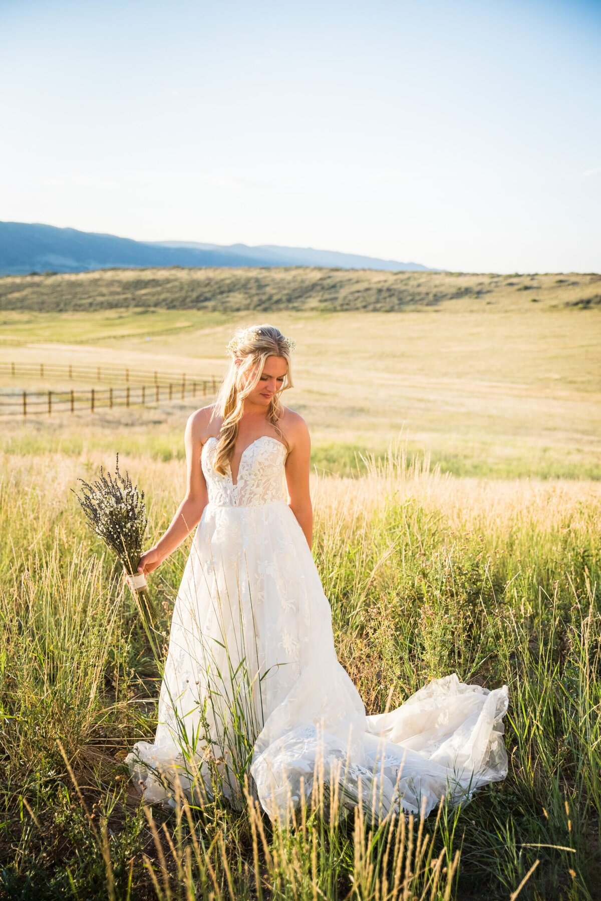 A bride holding her bouquet stands in a golden field and glances down at her wedding dress.