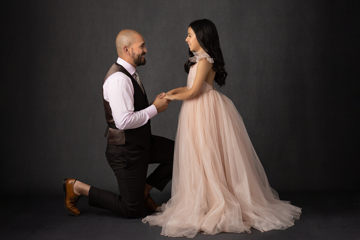 A Dallas family photographer captures a beautiful father-daughter moment as the father kneels and holds hands with his daughter, who is dressed in a soft pink gown.