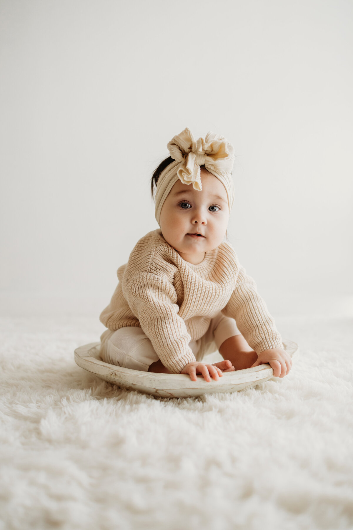 A happy eight month old baby sits in a wooden bowl during her milestone session in Denver while wearing a sweater and a large bow.