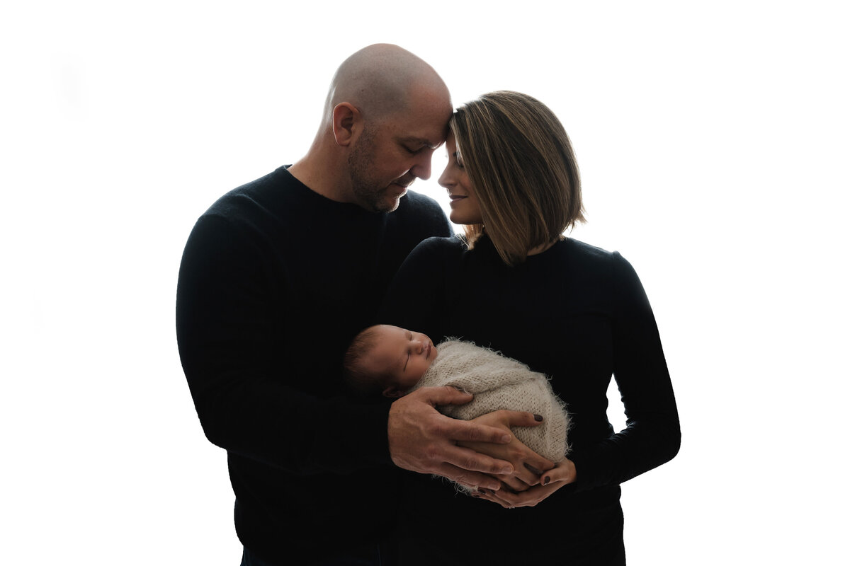 Mom and dad dressed in black pose cradling their newborn boy while closing their eyes and touching their foreheads and are lit from behind during their Pittsburgh newborn session.