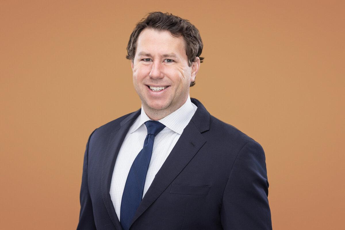 A confident male in a sleek navy suit and crisp white shirt exudes professionalism against a warm orange backdrop. His genuine smile captures the essence of modern business portraits, perfectly exemplifying the quality of Cincinnati headshots