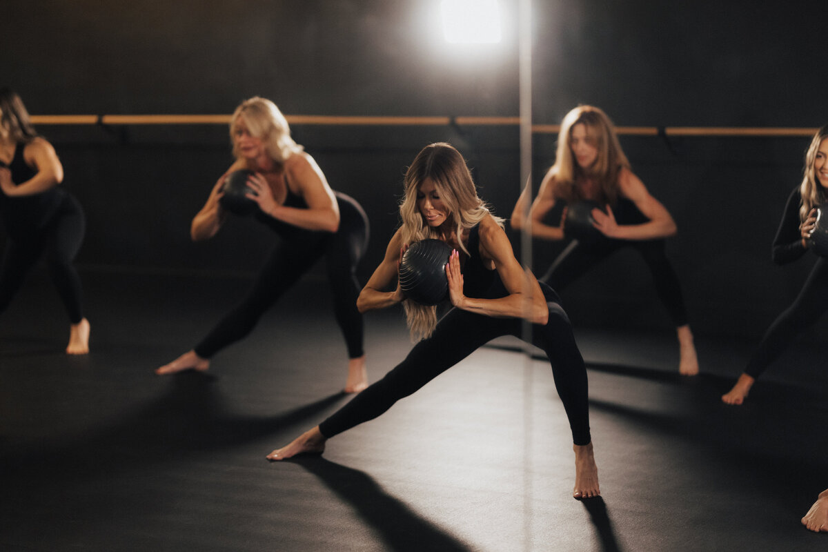 High-energy branding photo of women mid-movement in a dark fitness studio, using weighted balls during a group workout session — strong, bold, and empowering brand imagery.