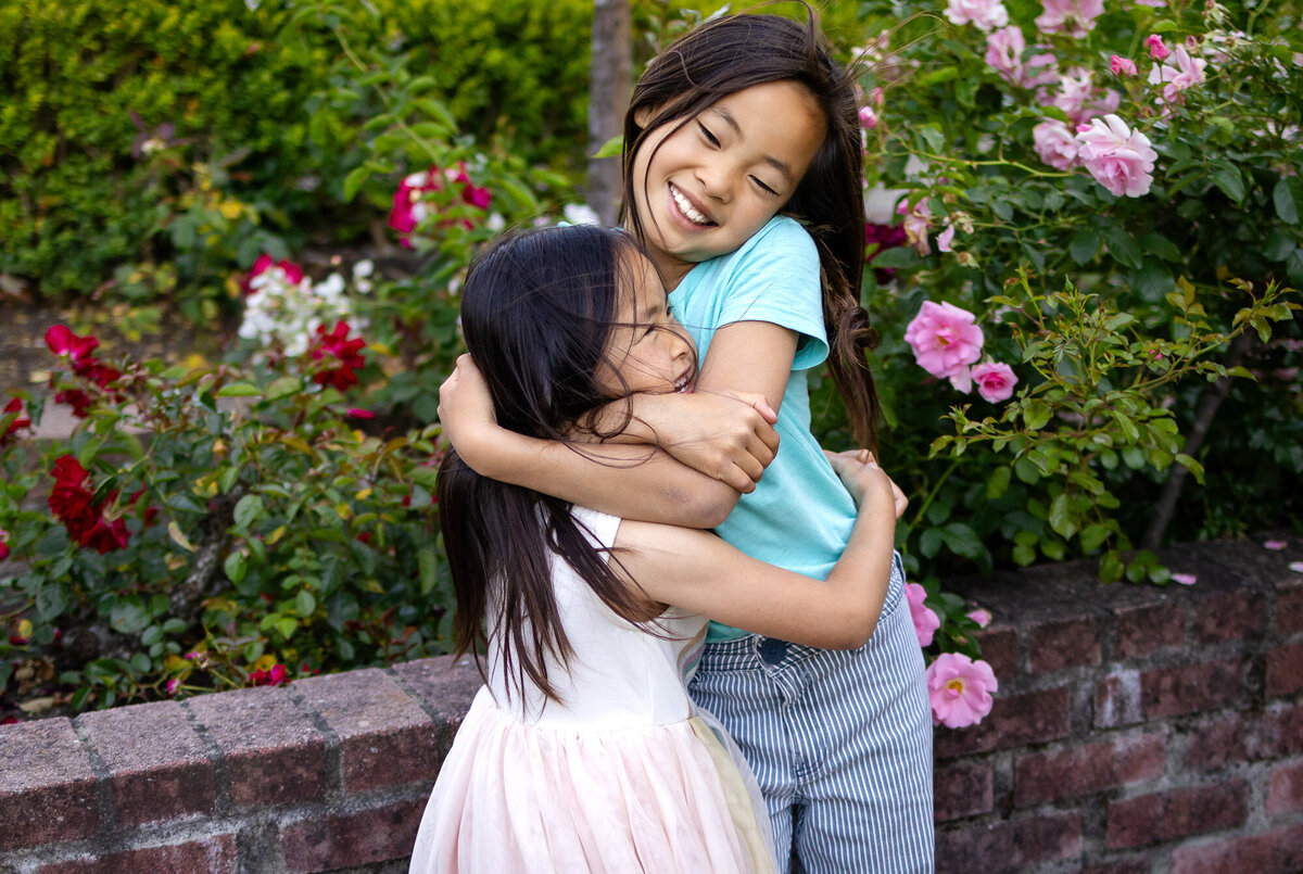 Big sister giving her younger sister a tight, loving hug in front of pink rose bushes – Bay Area Family Portfolio - Ellobelle Photography