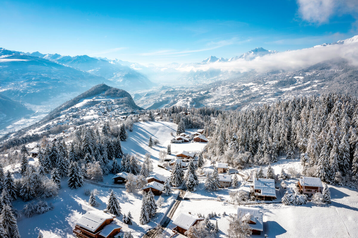 Aerial view of a snow-covered ski resort with slopes, surrounded by mountainous terrain. Various lodges and buildings are nestled below, interconnected by roads. Pine trees dot the landscape under a clear sky.