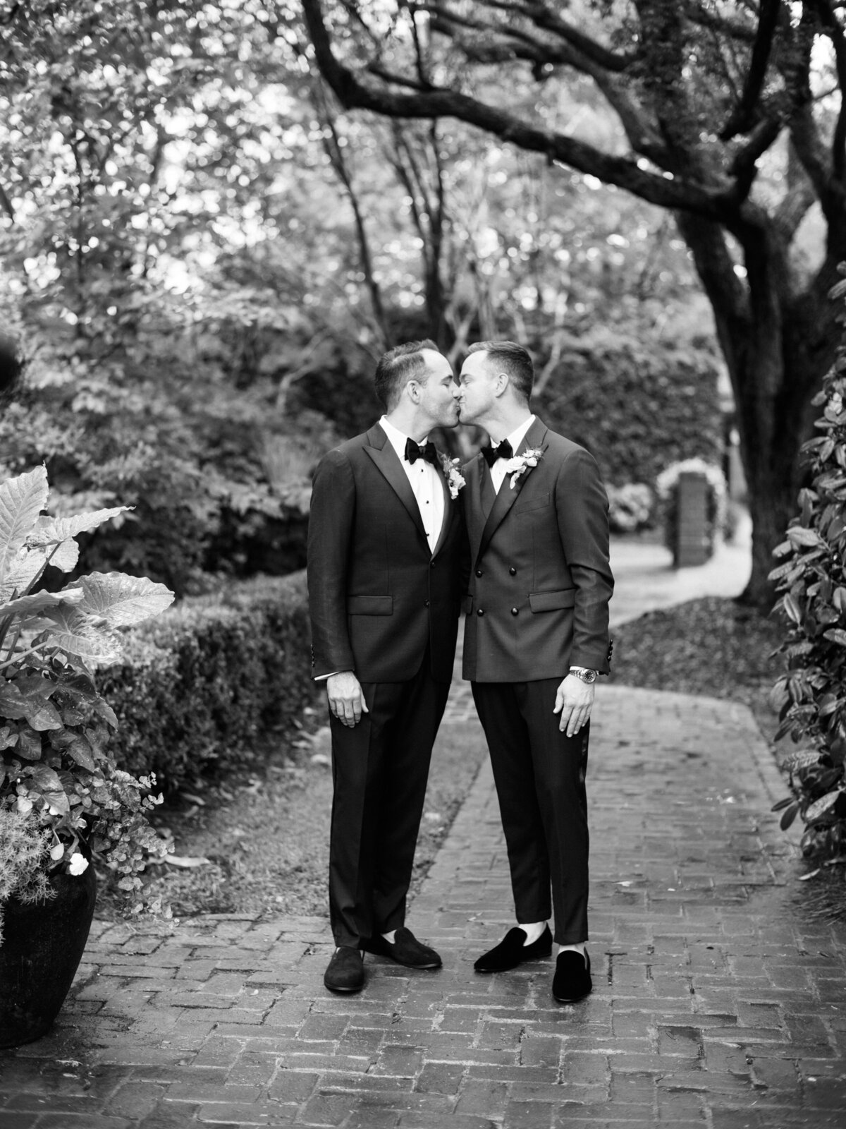 A romantic moment captured between two grooms sharing a kiss, framed by lush greenery and a charming pathway, celebrating love and commitment on their wedding day in Charleston, SC.