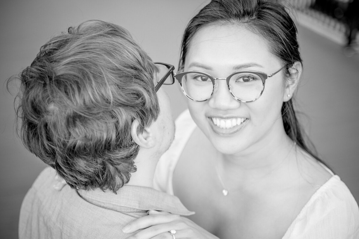 Black-and-white-top-down-angle-of-a-smiling-Asian-female-wearing-glasses-during-her-engagement-session-at-the-University-City-Boardwalk-by-Charlotte-wedding-photographers-DeLong-Photography