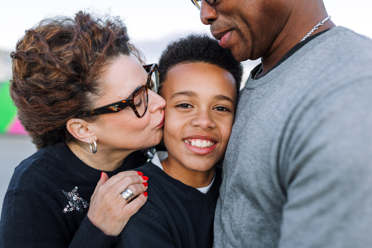 mom kissing 11-year-old son as he leans on his dad's chest and looks at camera smiling