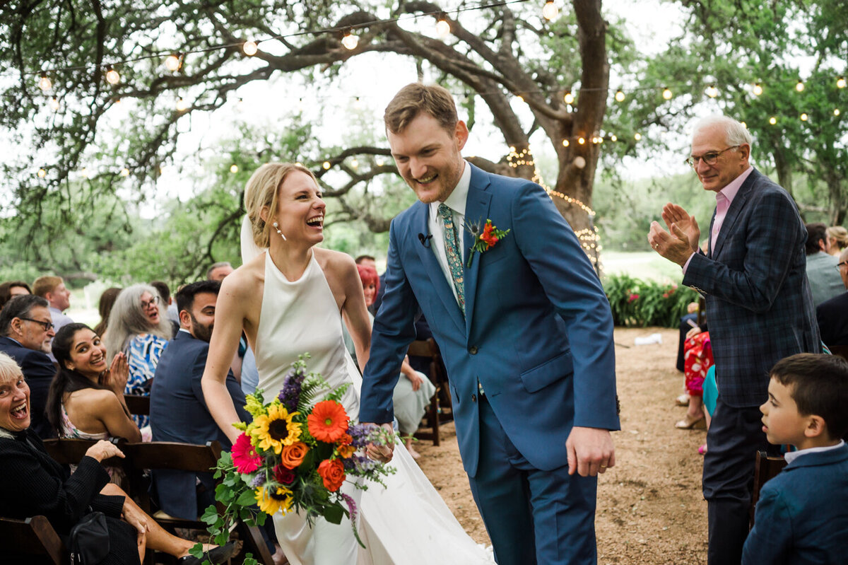 A joyful bride and groom walk down an outdoor aisle under tree lights, smiling and laughing. Guests clap and cheer, surrounded by greenery and flowers.