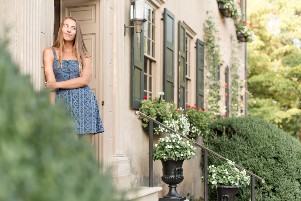 Senior girl leaning against historic house in Lancaster, Pennsylvania with left hand on right arm looking left.