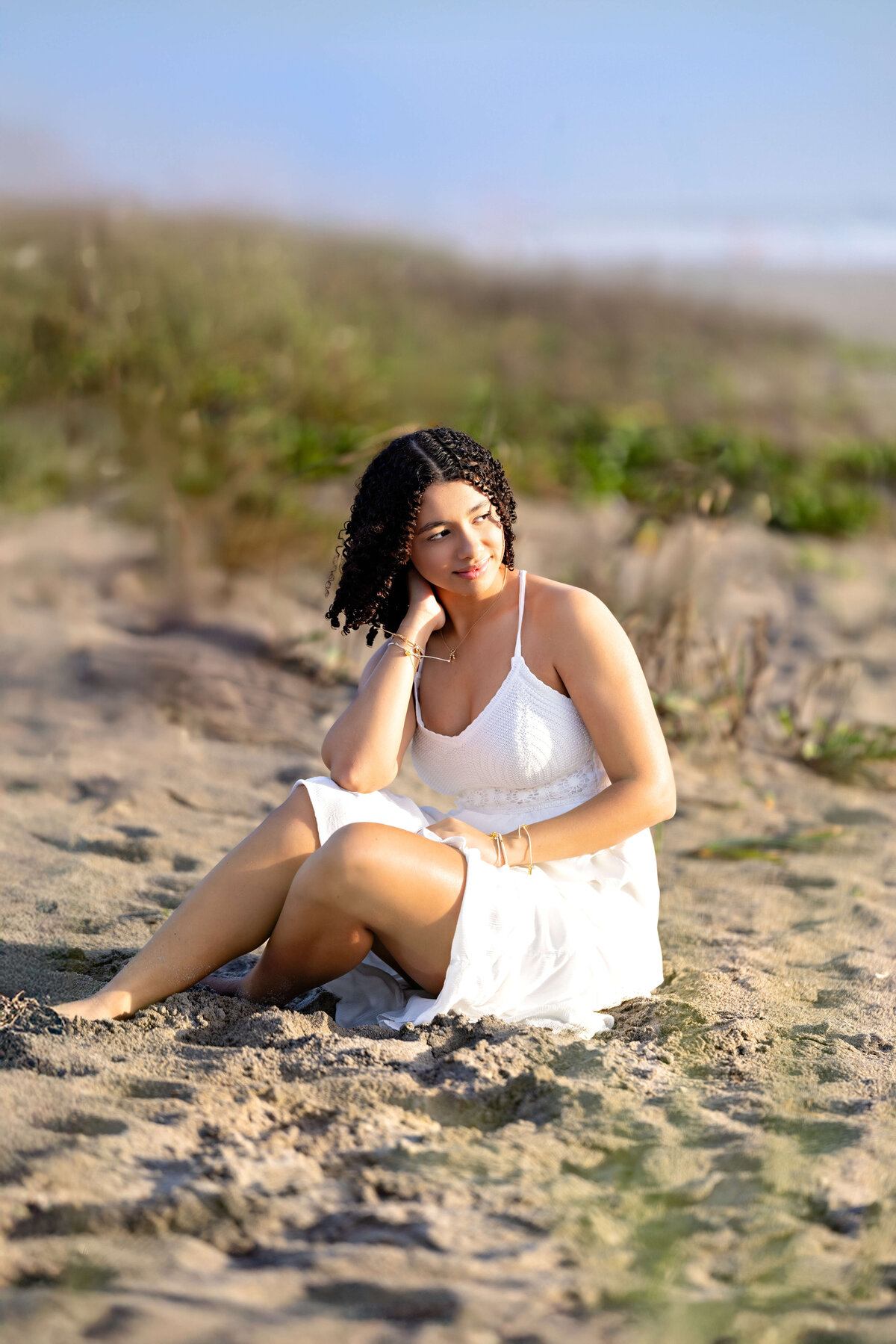 Cocoa Beach senior session featuring a girl standing in the sand looking off into the distance, sunlight by direct light with beach sand and grass around her.